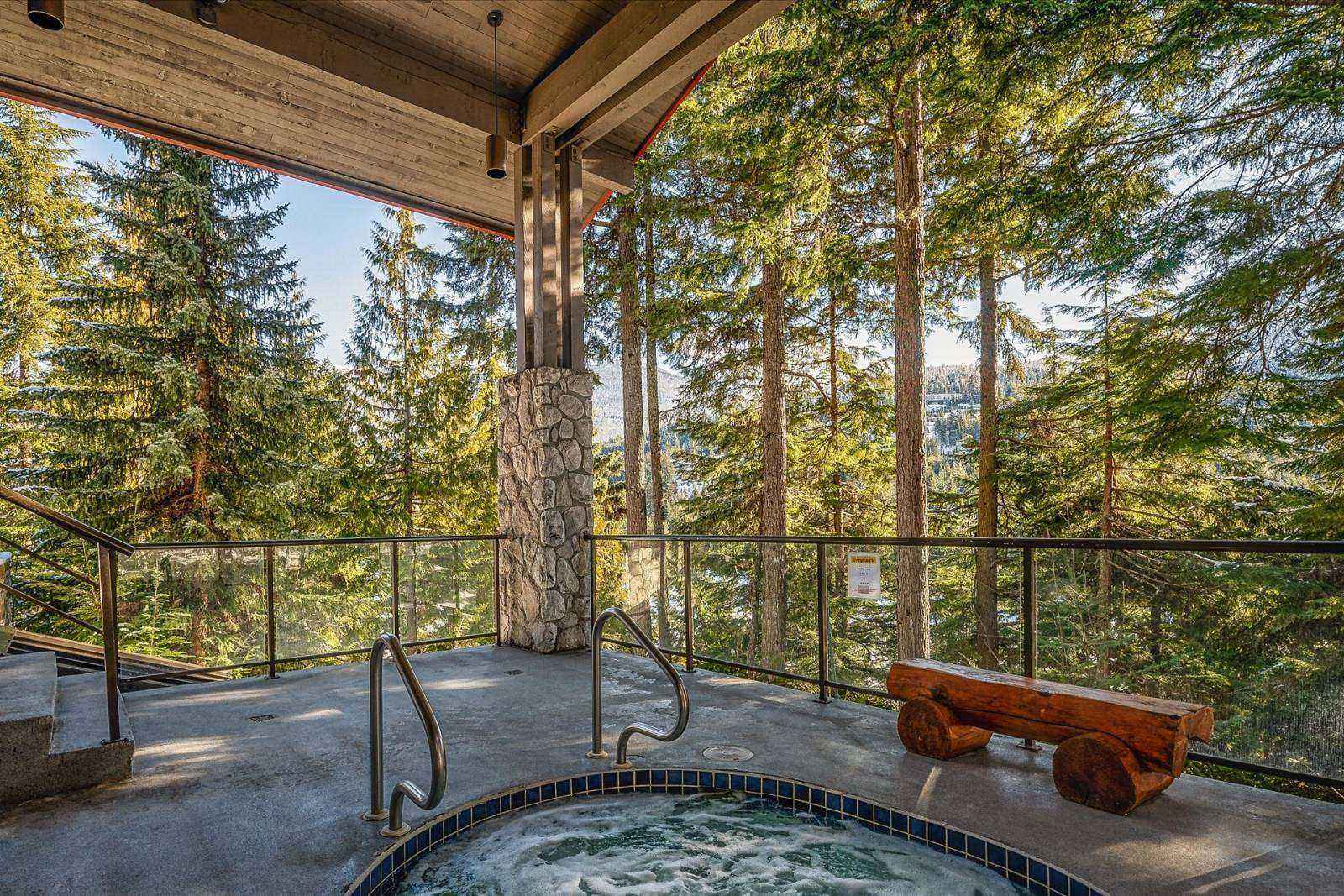 The communal hot tub on a covered deck, surrounded by tall evergreens with mountain views beyond.